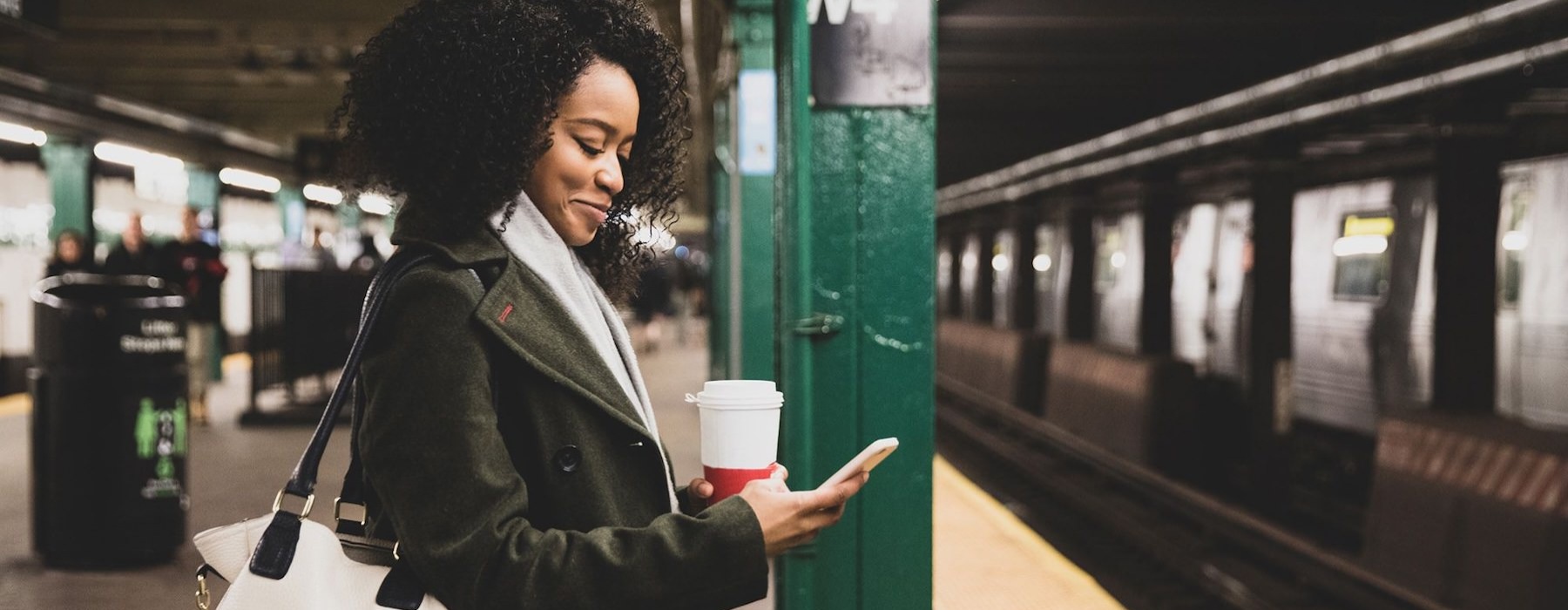 woman with a cup of coffee texts on subway station platform